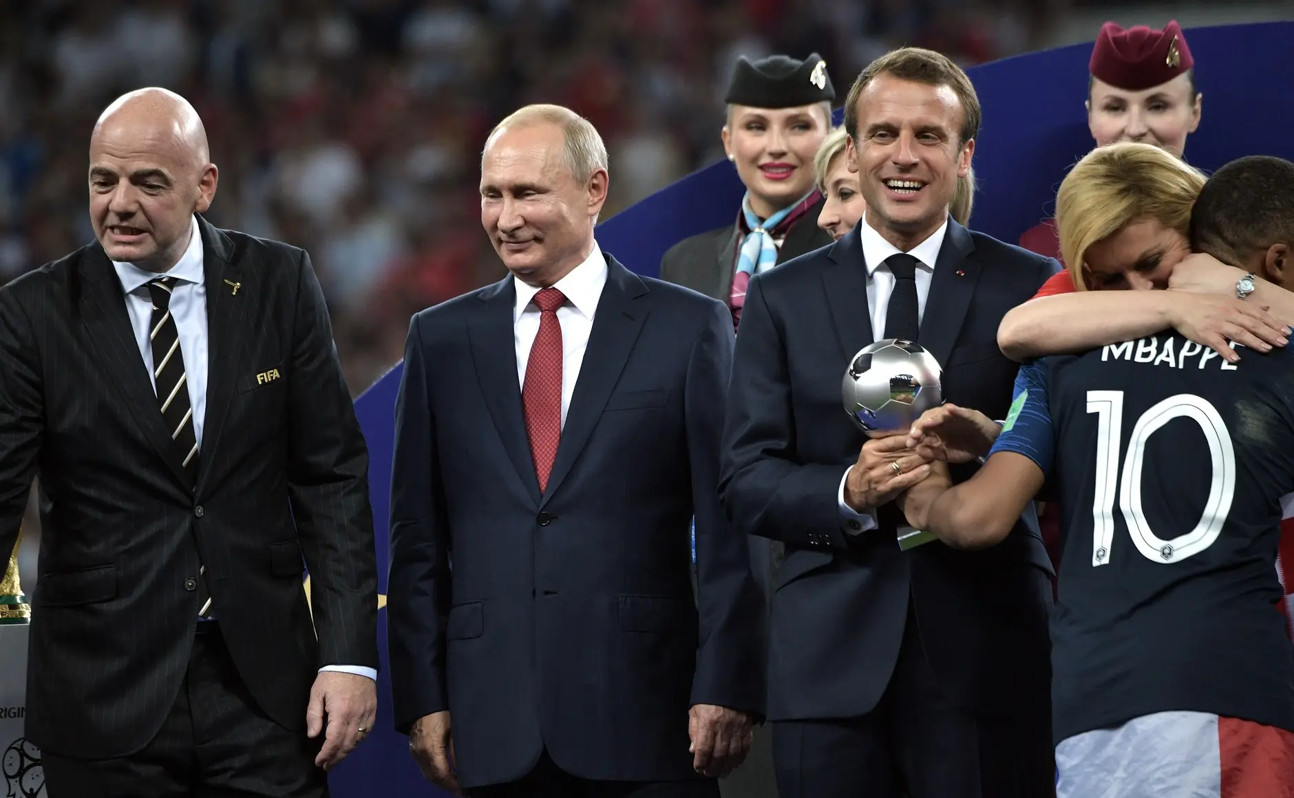 Kylian Mbappé de costas, vestindo a camisa 10 da França, recebendo um prêmio do presidente Emmanuel Macron.